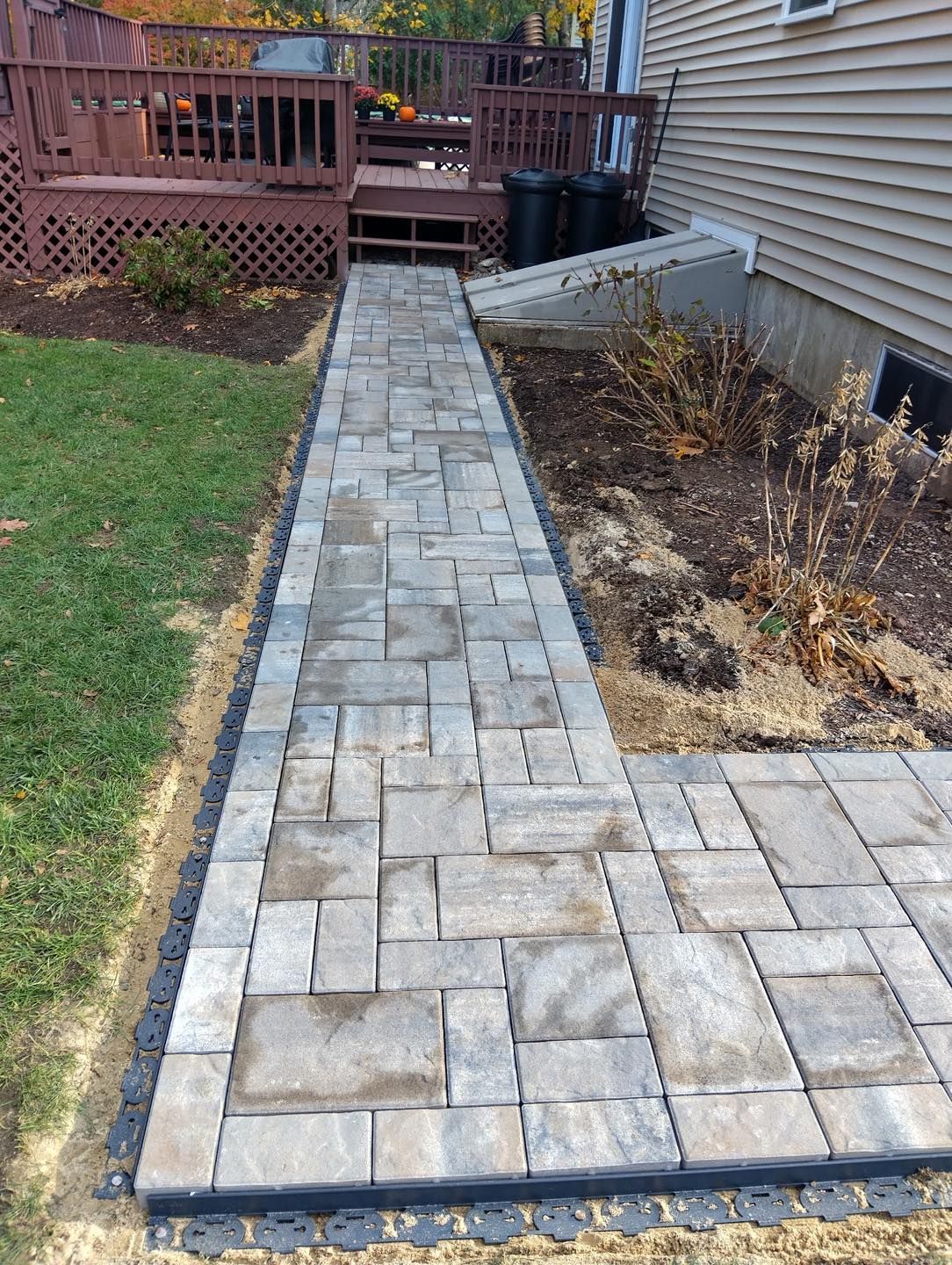 Stone paver walkway along a house, bordered by lawn and a mulch bed, leading to a wooden deck.
