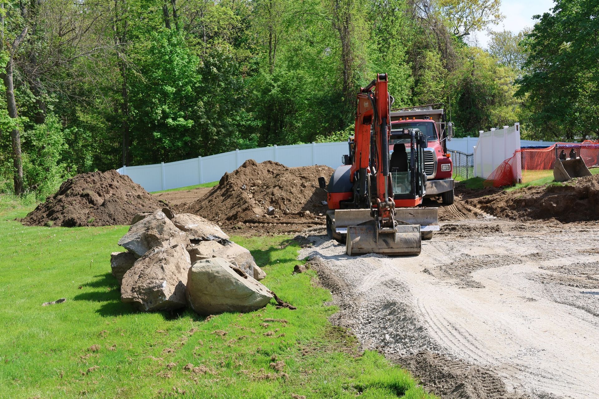 Excavator on a gravel road beside dirt piles and large rocks, with green grass and trees nearby