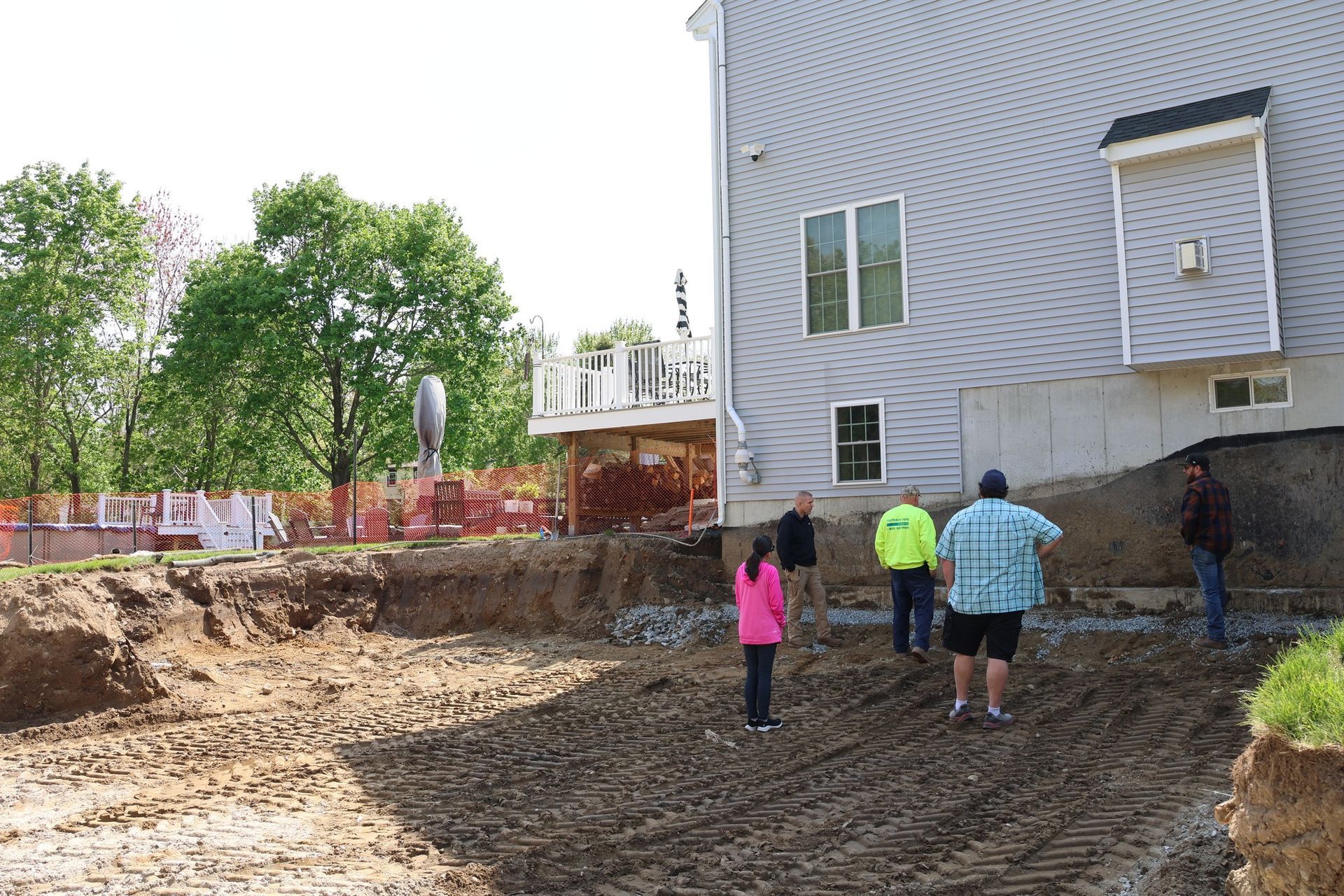 Three people stand in a muddy backyard beside a gray house and a fenced yard.