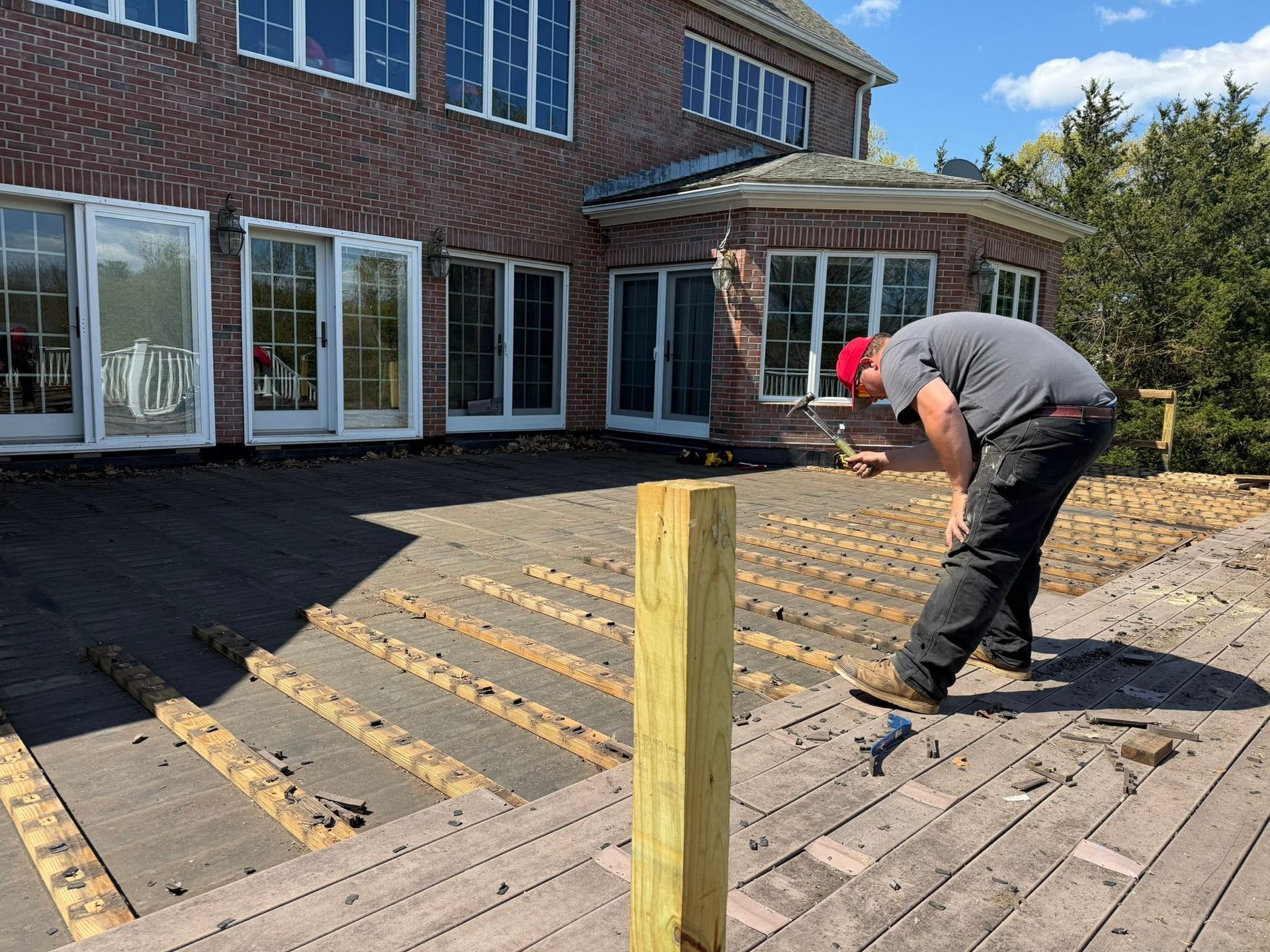 Worker sanding a wooden deck beside a brick house on a sunny day