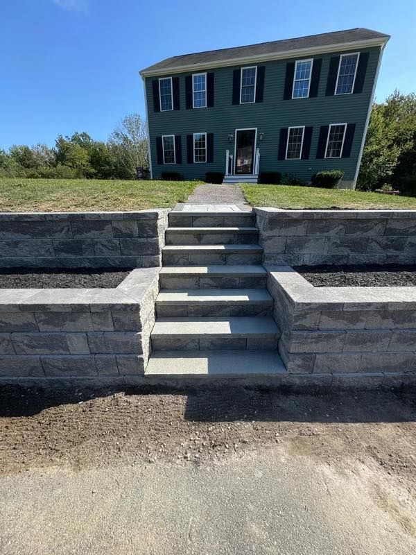 Stone staircase leading up to a two-story dark house with black shutters on a grassy hill under a blue sky