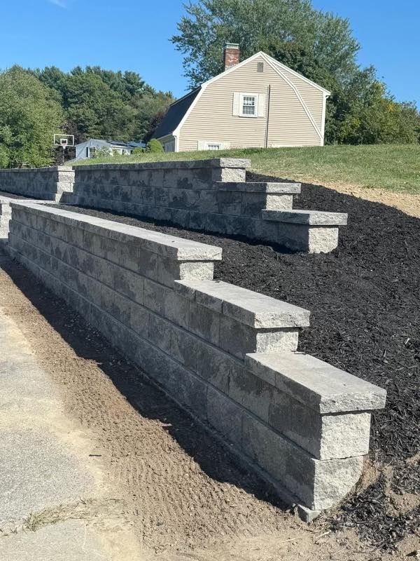 Tiered gray stone retaining wall beside a driveway, with black mulch and a beige house on a grassy slope.