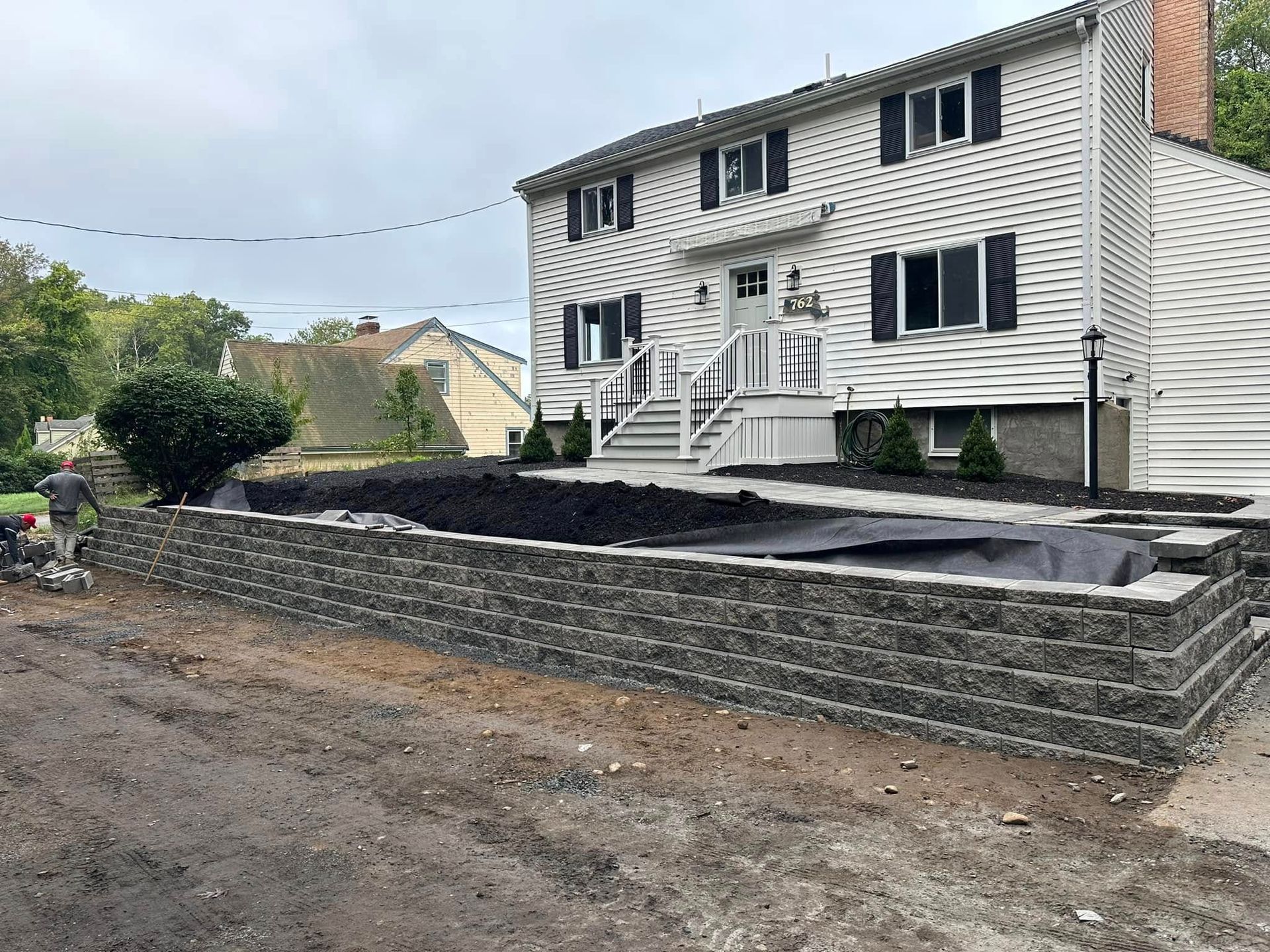 Stone retaining wall and freshly graded backyard beside a white house with stairs and black shutters