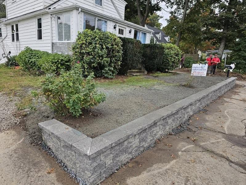 White house with gravel front yard, low concrete border, bushes, and two people near a sign.