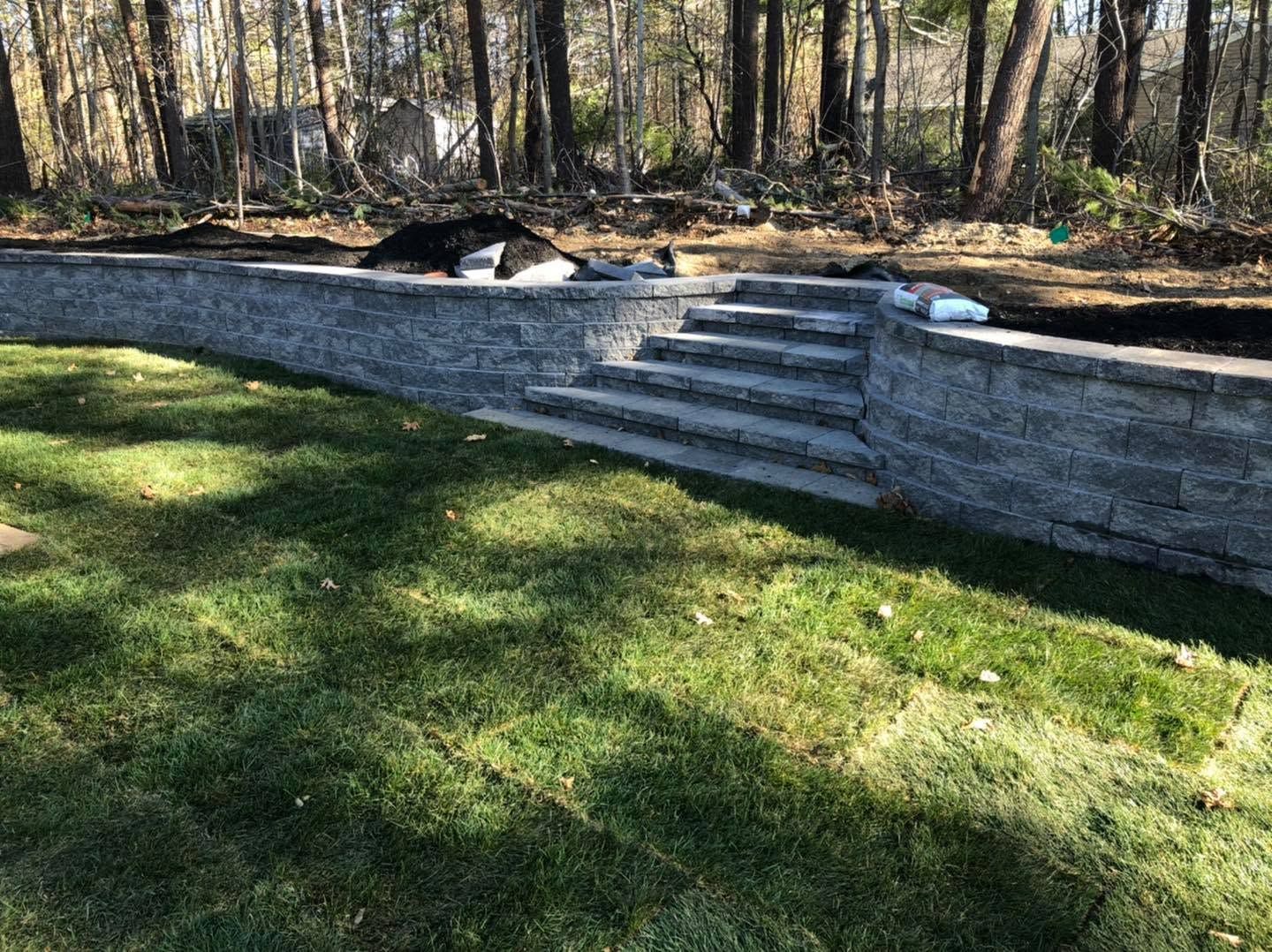 Stone retaining wall with steps leading up to a wooded yard, beside a grassy lawn.