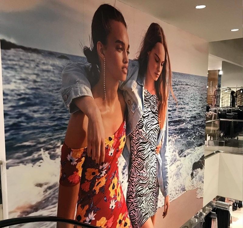 Models in summer dresses, one with arm around the other, in front of a beach background.