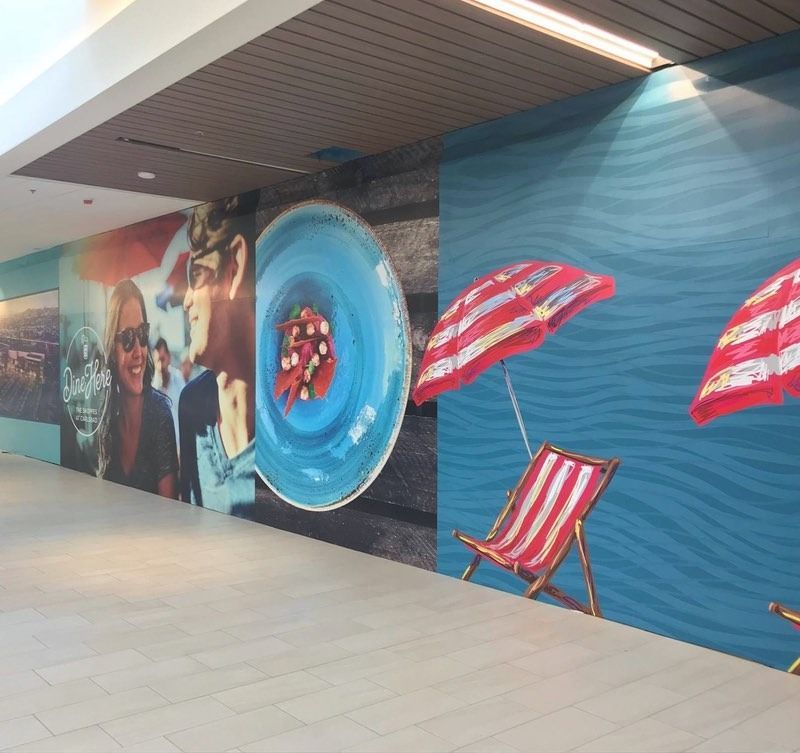 Mall wall mural with beach umbrellas, lounge chairs, and a person looking through a blue circle.