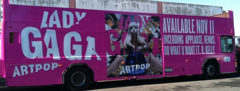 Pink bus advertisement for Lady Gaga's ARTPOP album, featuring an image of her and text stating availability.