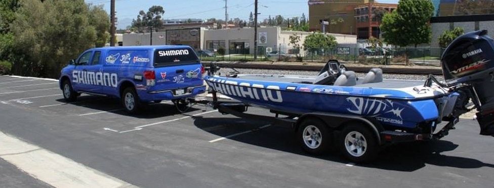 Blue Shimano truck towing a boat on a trailer, parked in a lot.