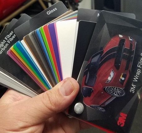 Hand holding a color swatch fan of 3M vinyl car wrap samples. Glossy colors and carbon fiber options.