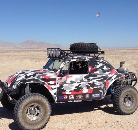 Off-road modified VW Beetle in desert setting, camo paint, spare tire on roof rack.