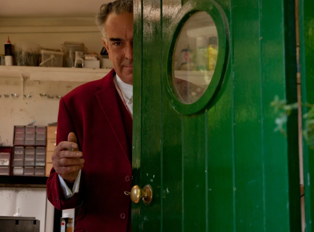 Man in red suit peers from green door with round window.