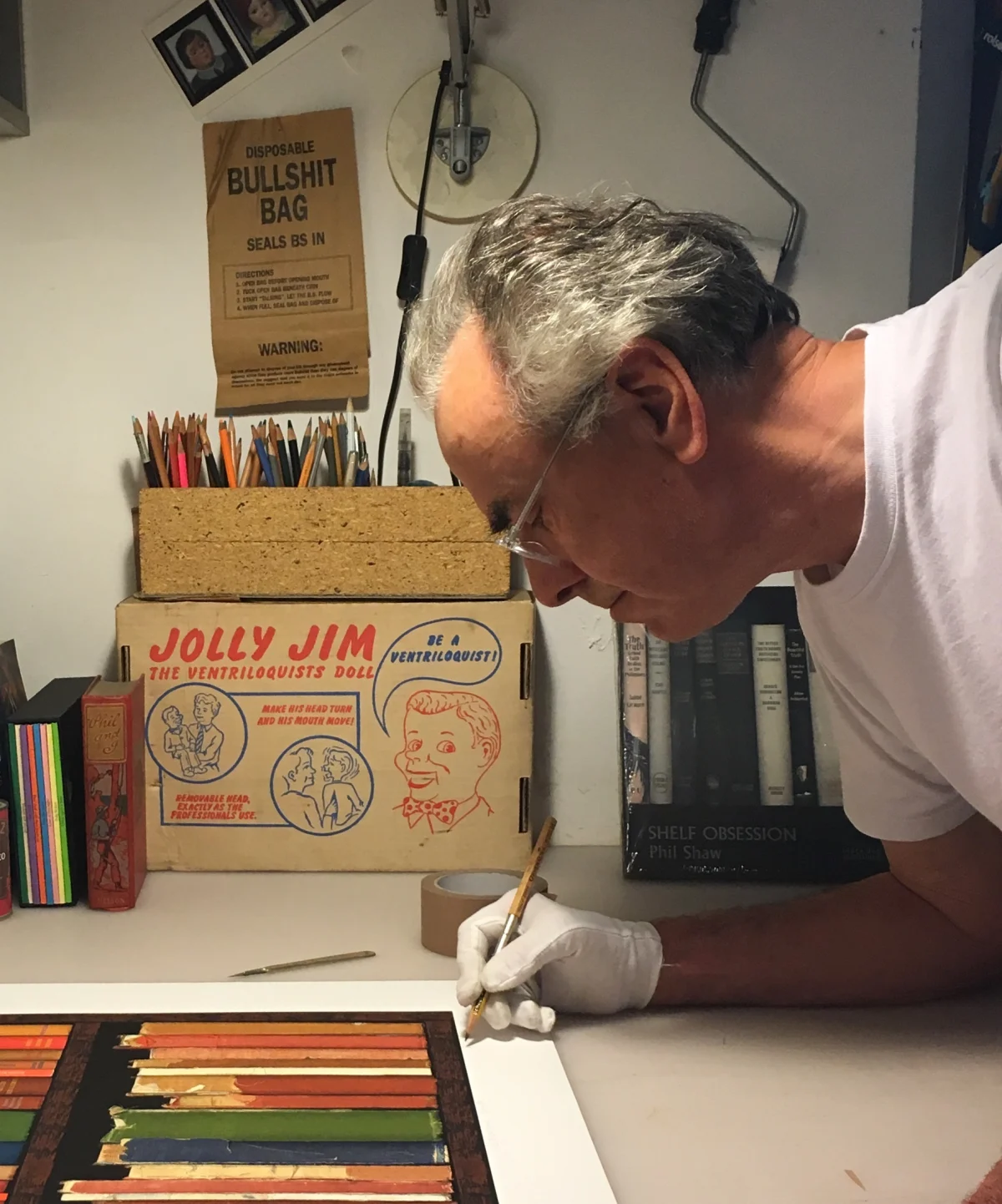 Man in glasses wearing a white glove working at a table with colored pencils, a box labeled