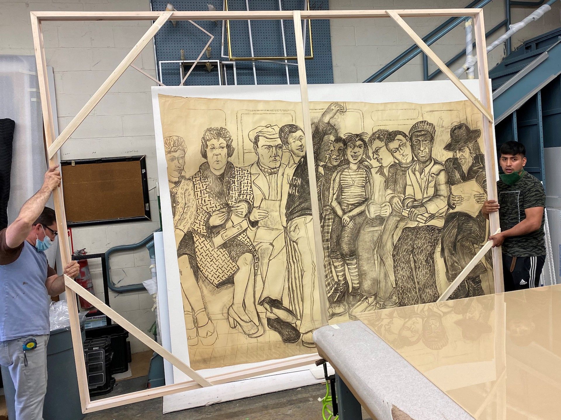 Two people frame a large black and white drawing of many figures, likely a mural, in a workshop.