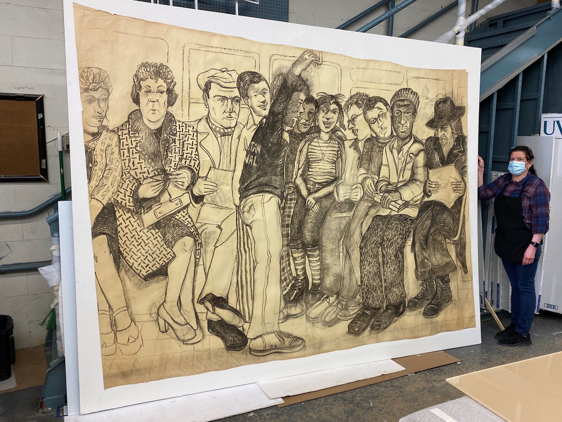 Large charcoal drawing of people; a woman in a mask and apron holds it against a warehouse wall.