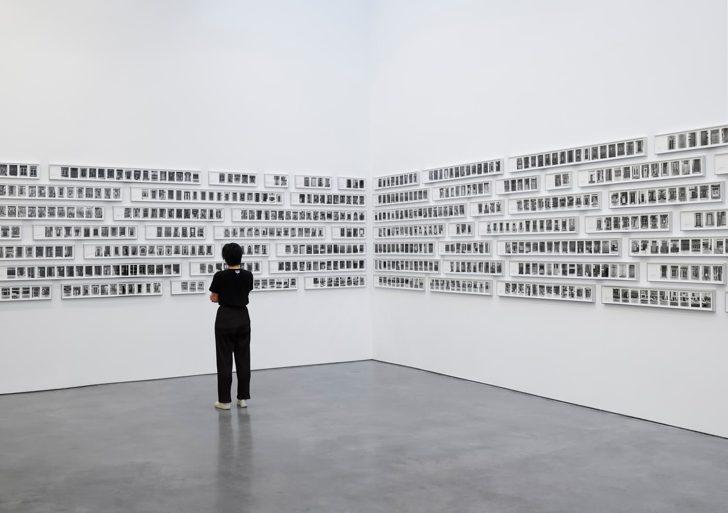 Person viewing rows of framed, text-based artwork on white gallery walls.