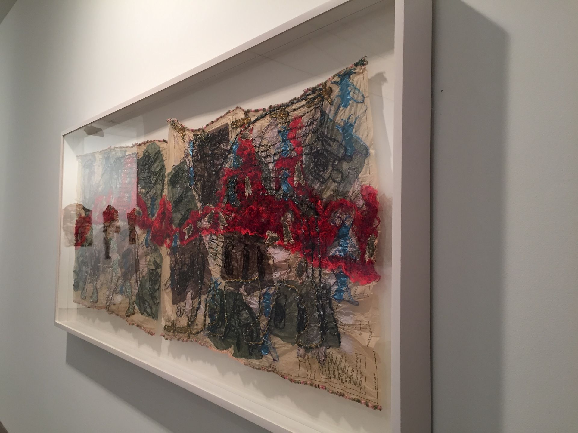 Artwork in a white frame on a white wall. The art features red, blue, and brown abstract shapes.