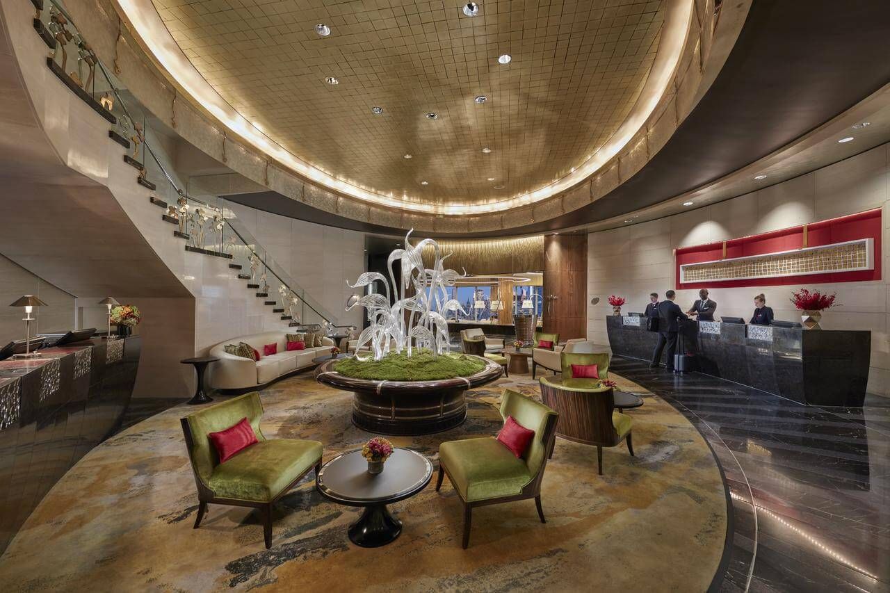 Hotel lobby with staircase, reception desk, seating area, and decorative fountain under a gold-toned ceiling.