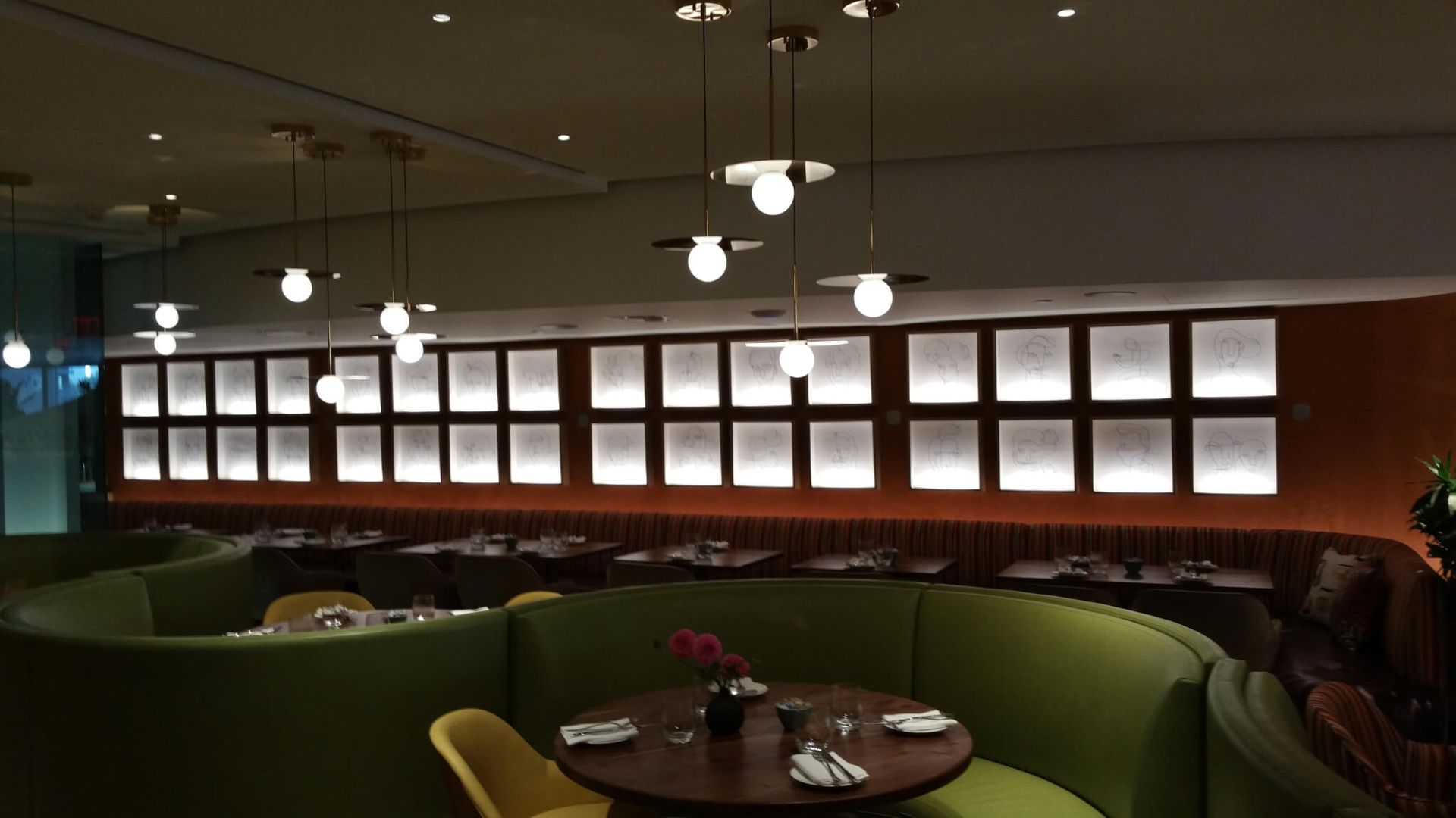 Restaurant interior with green curved booths, round tables, and pendant lights. A wall features backlit square panels.