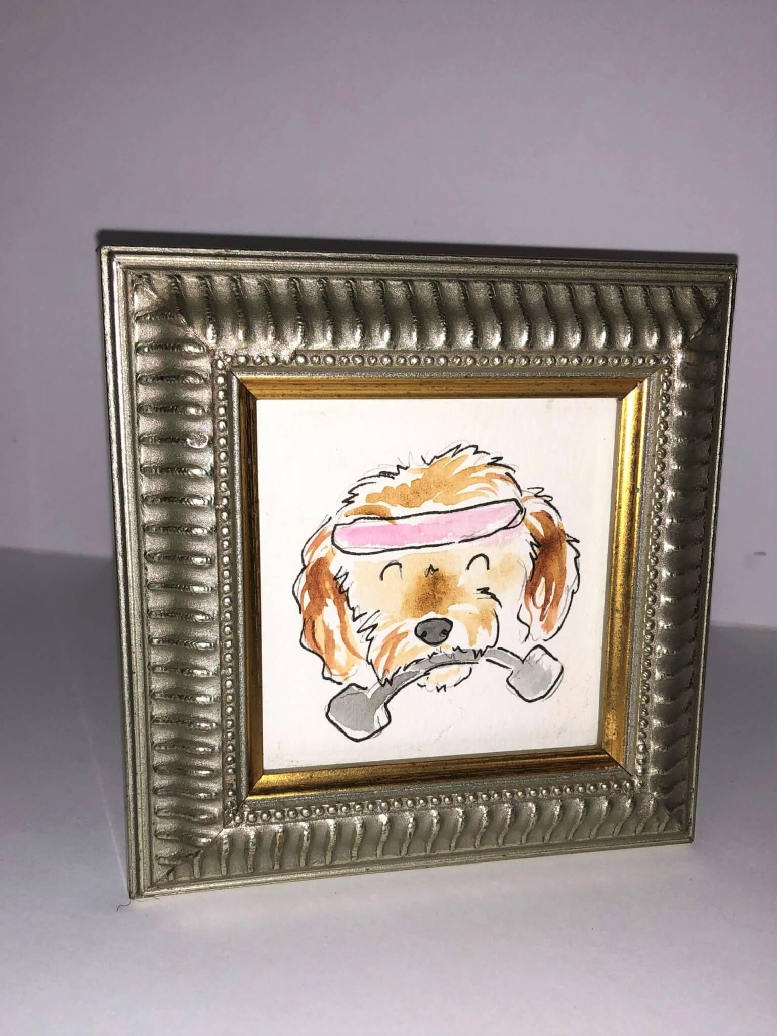 Watercolor of a dog with a headband lifting weights, framed in silver and gold.