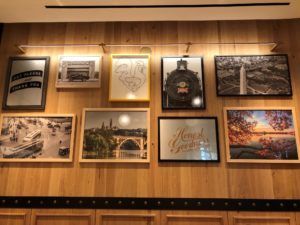 Framed art display on wood panel wall: various photographs and text art with brass lighting and trim.