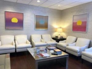 Awaiting room with white chairs, three abstract paintings, and a coffee table with reading material.