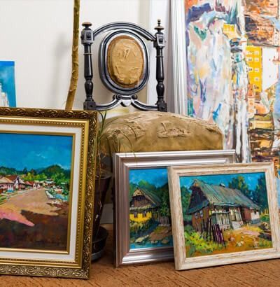 Paintings stacked near a decorative chair, with landscape scenes in colorful frames.