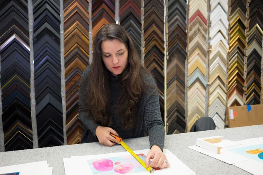 Woman measures artwork with a tape measure, surrounded by a wall of picture frames.