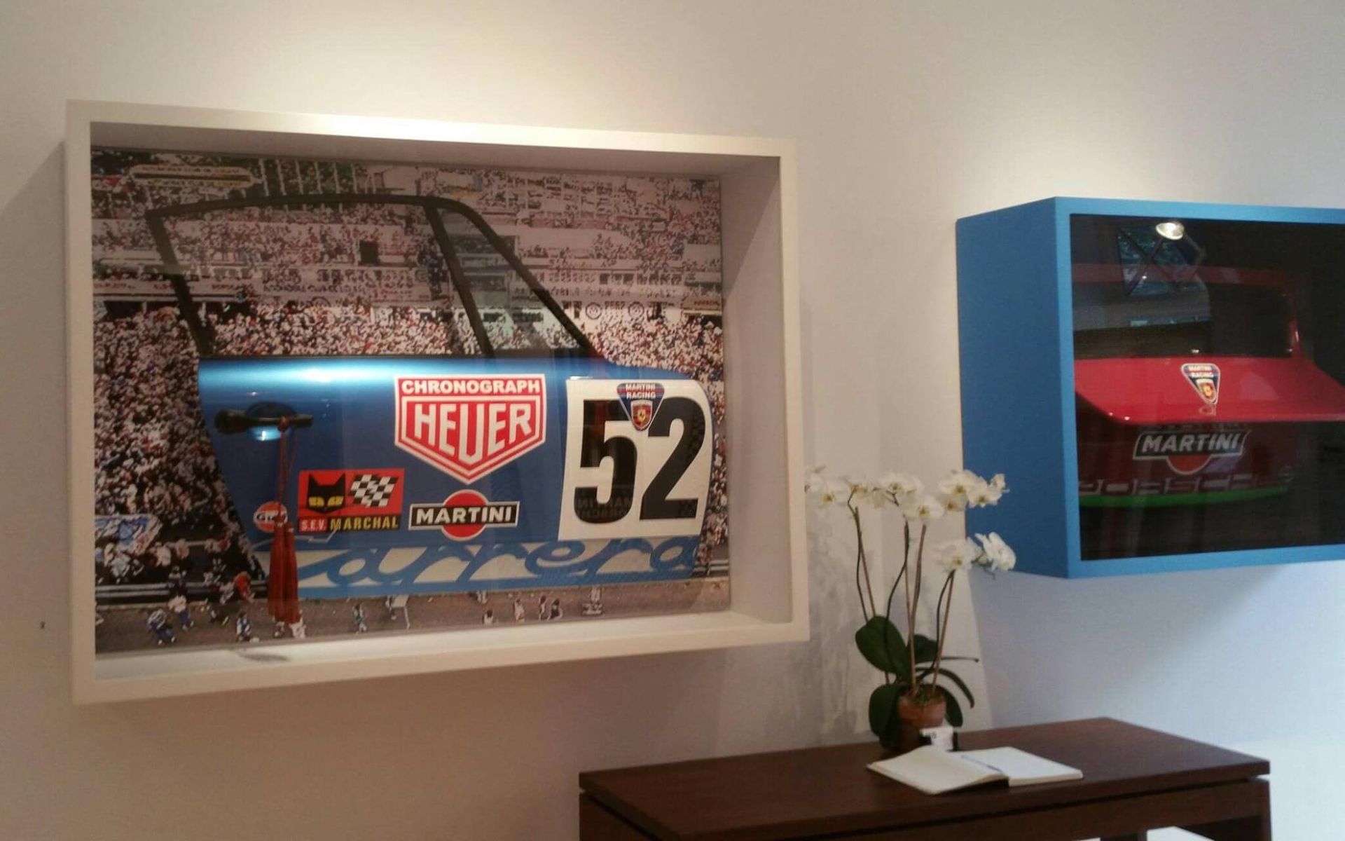 Art gallery display: a framed Porsche car door with a crowd background, and a blue display case with a red car model.