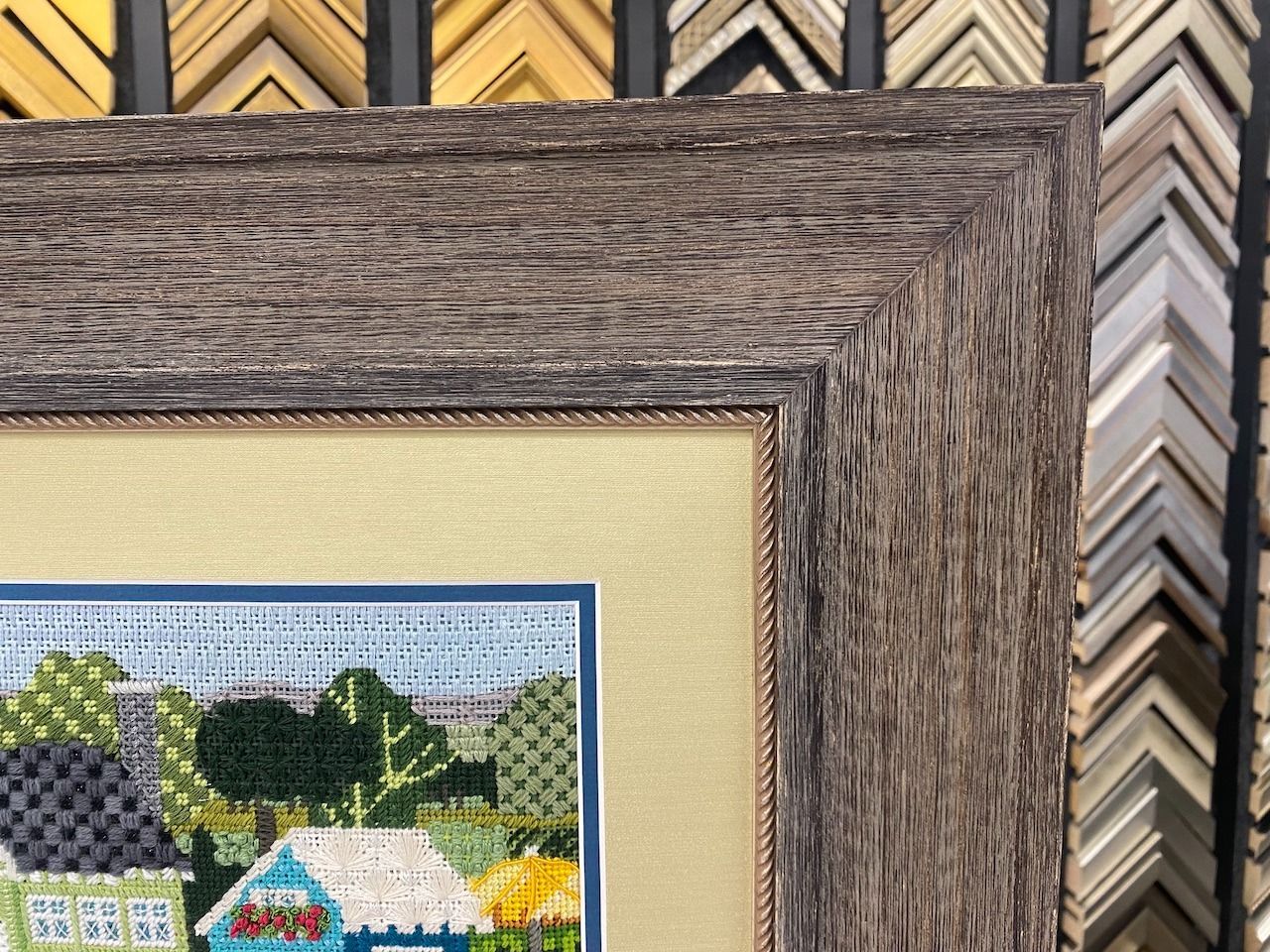 Close-up of framed cross-stitch art; rustic gray wooden frame surrounds the artwork.