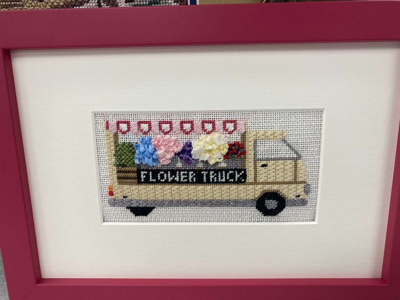 Cross-stitched flower truck framed in pink. Beige truck filled with colorful flowers. 