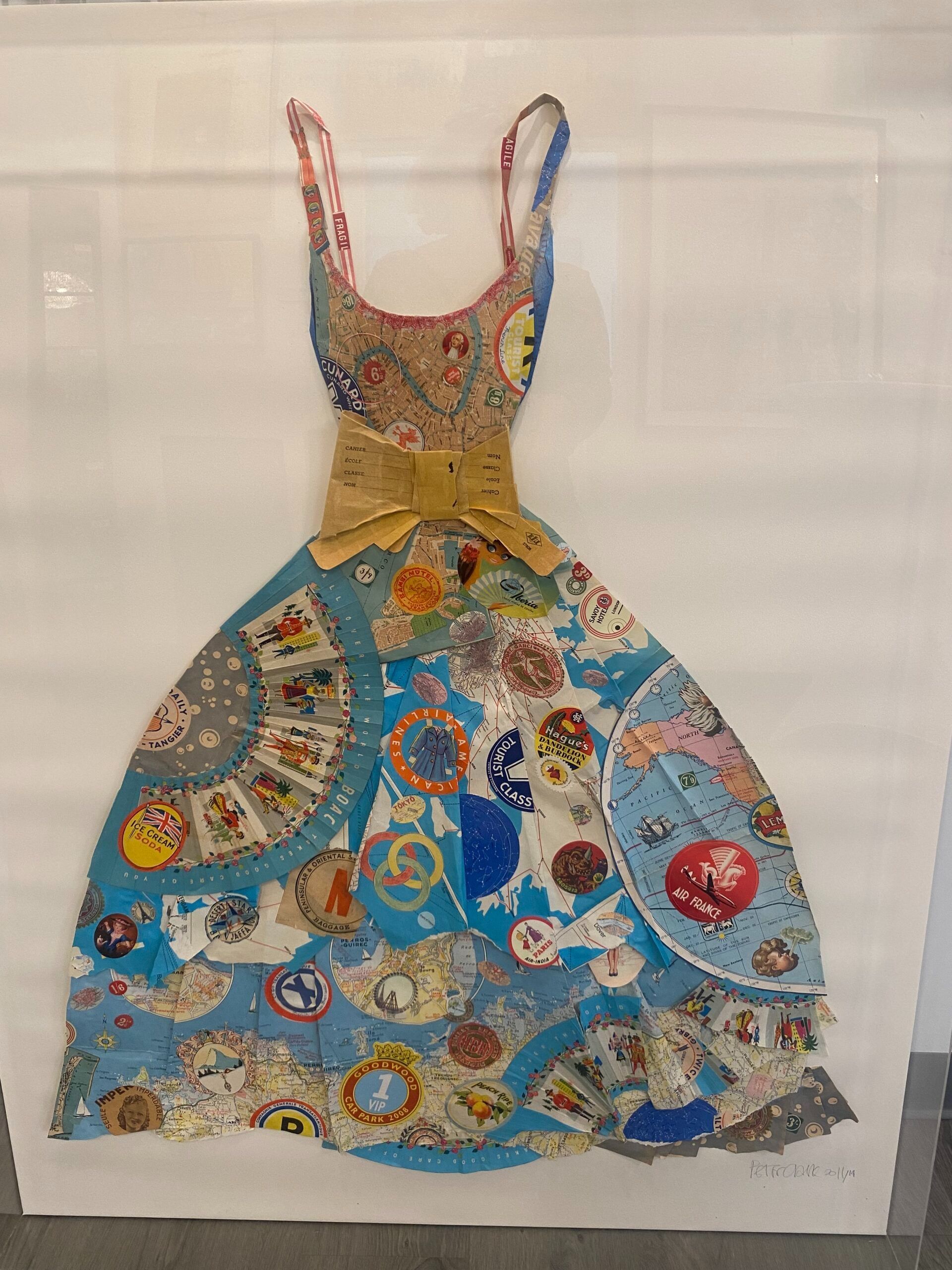 Dress-shaped artwork made of various circular objects, primarily blue. Features a yellow bow and thin straps, set against a white background.