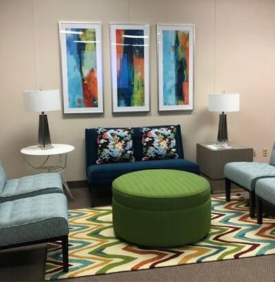 Lounge area with teal furniture, colorful artwork, a green ottoman, and a patterned rug.