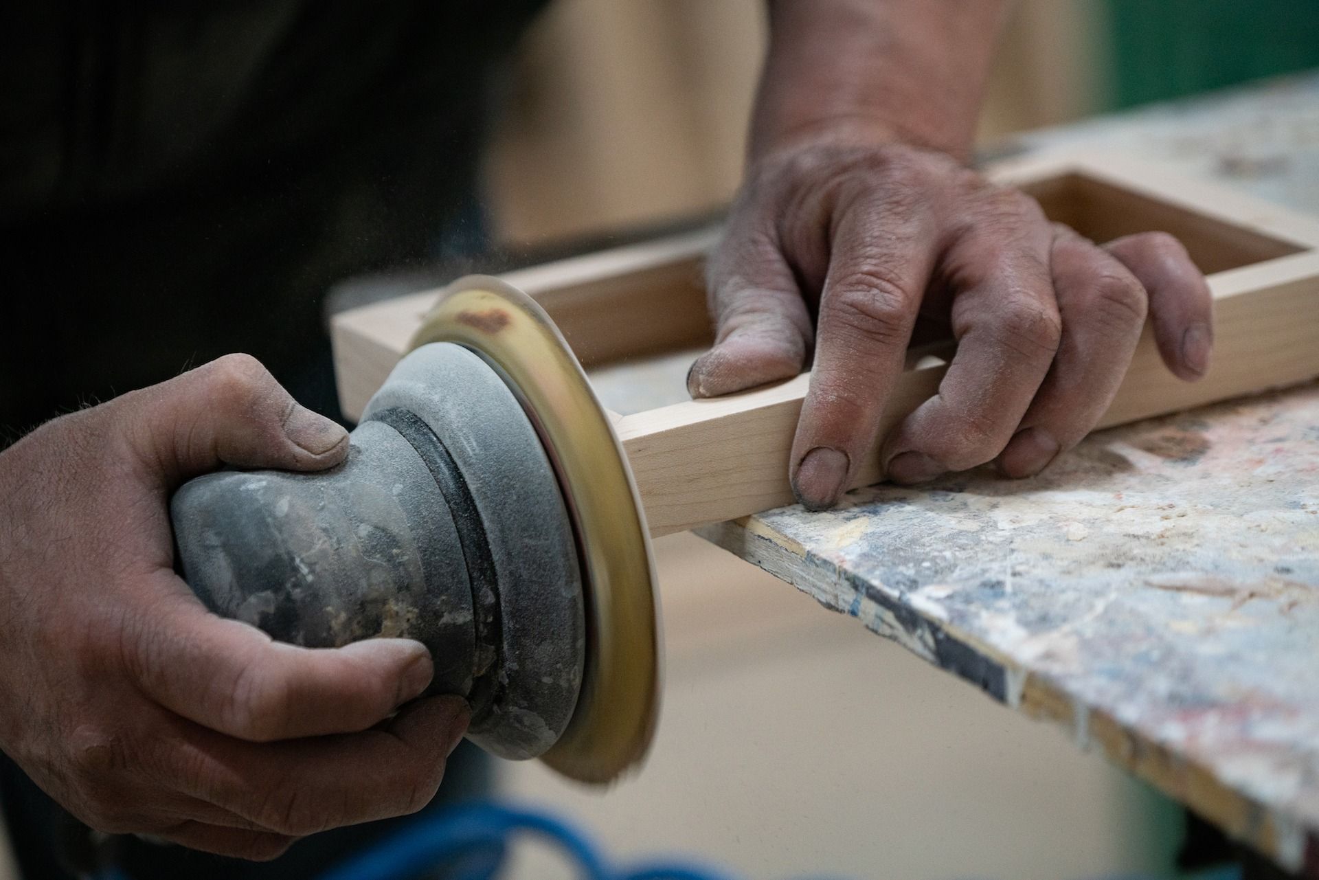 Hands sanding wooden frame with a power sander on a workbench.