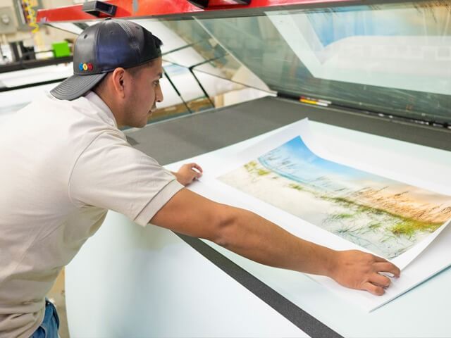 Man in cap examines a large, colorful print of a landscape on a light table.