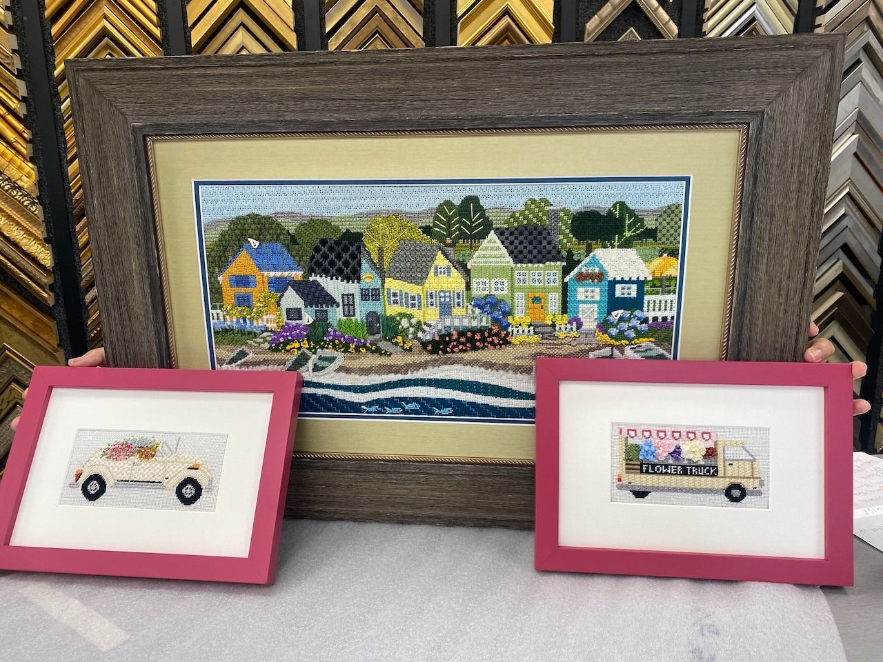 Framed needlepoint art: a beach town scene and two food truck pieces, all in pink and brown frames.