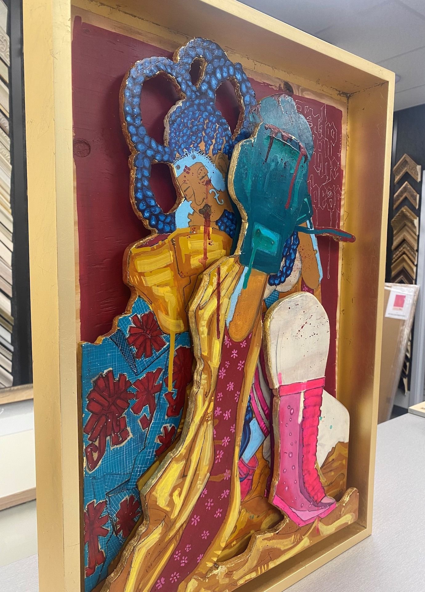 3D artwork with layered figures in bright colors framed in wood, set against a red background.