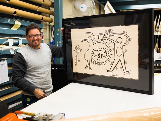 Man holding a framed Keith Haring drawing; a white figure on beige background with black frame.