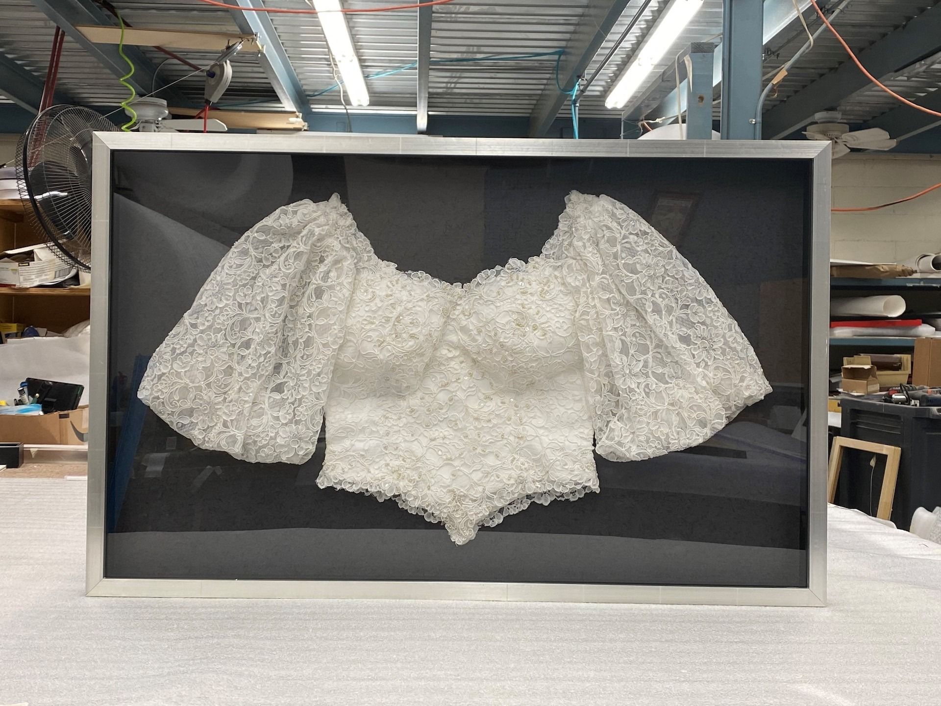 White lace bodice with large sleeves framed in a shadow box against a dark background.