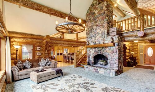 Rustic living room with stone fireplace, wooden beams, loft railing, and patterned sofa under a chandelier.