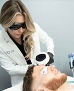 Esthetician wearing sunglasses performs a facial treatment on a reclining man in a clinic