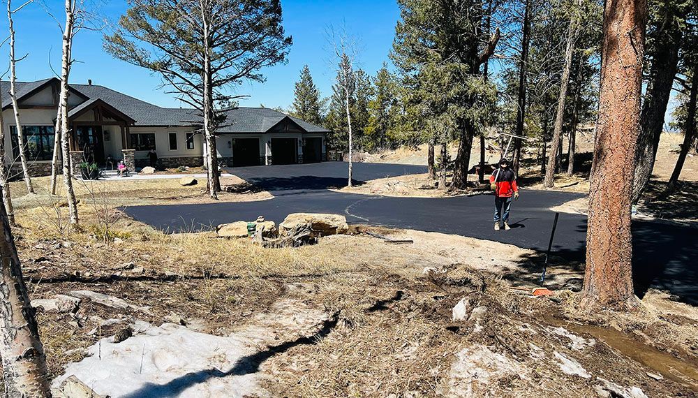 a man is walking down a dirt road in front of a house surrounded by trees.