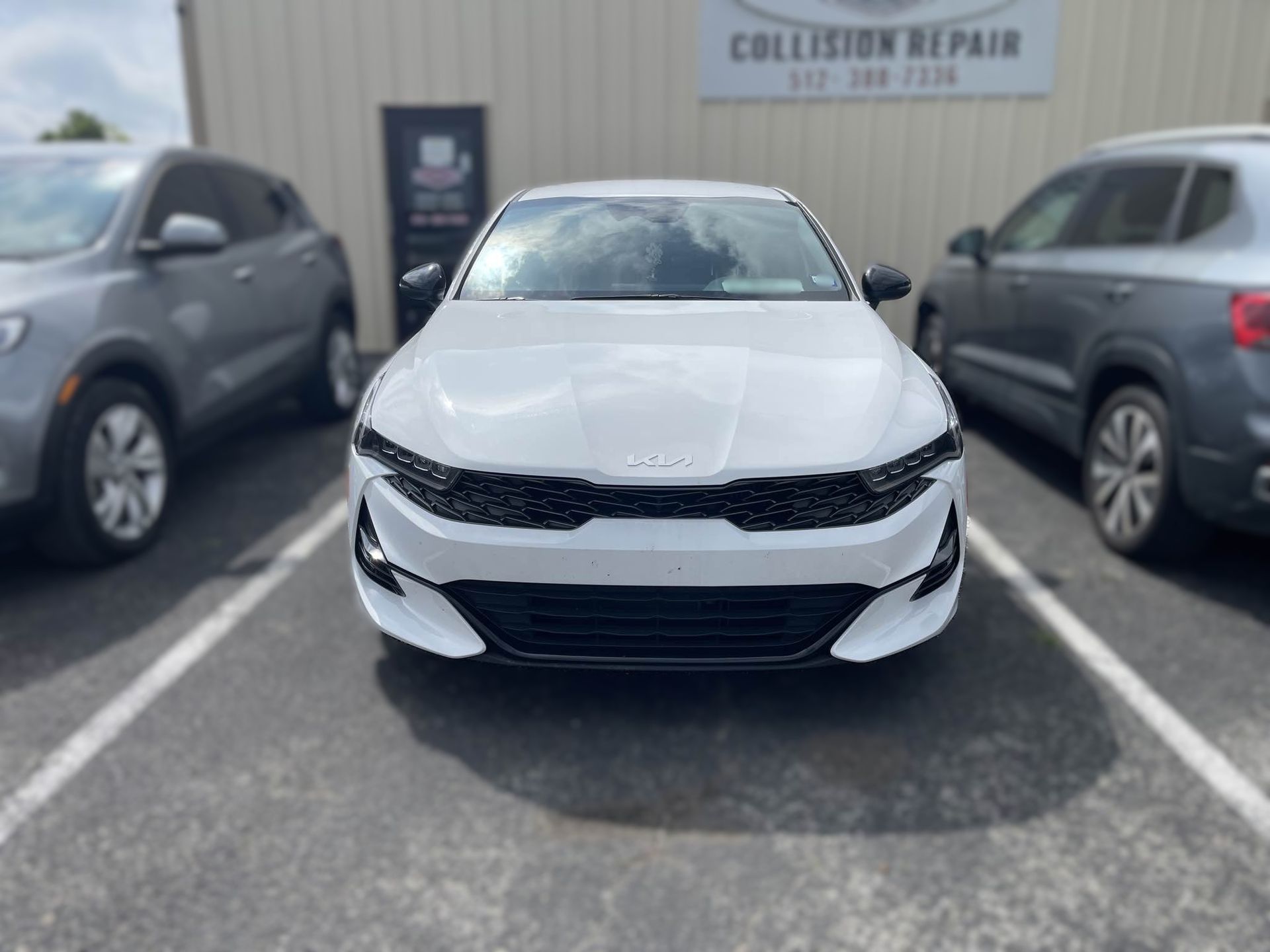 White Kia sedan parked in front of a collision repair shop, flanked by two other cars.