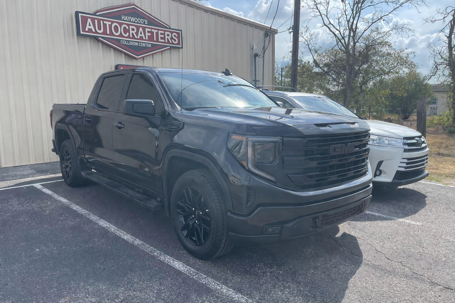 A black GMC truck is parked in front of the  Autocrafters shop, with a white car next to it.