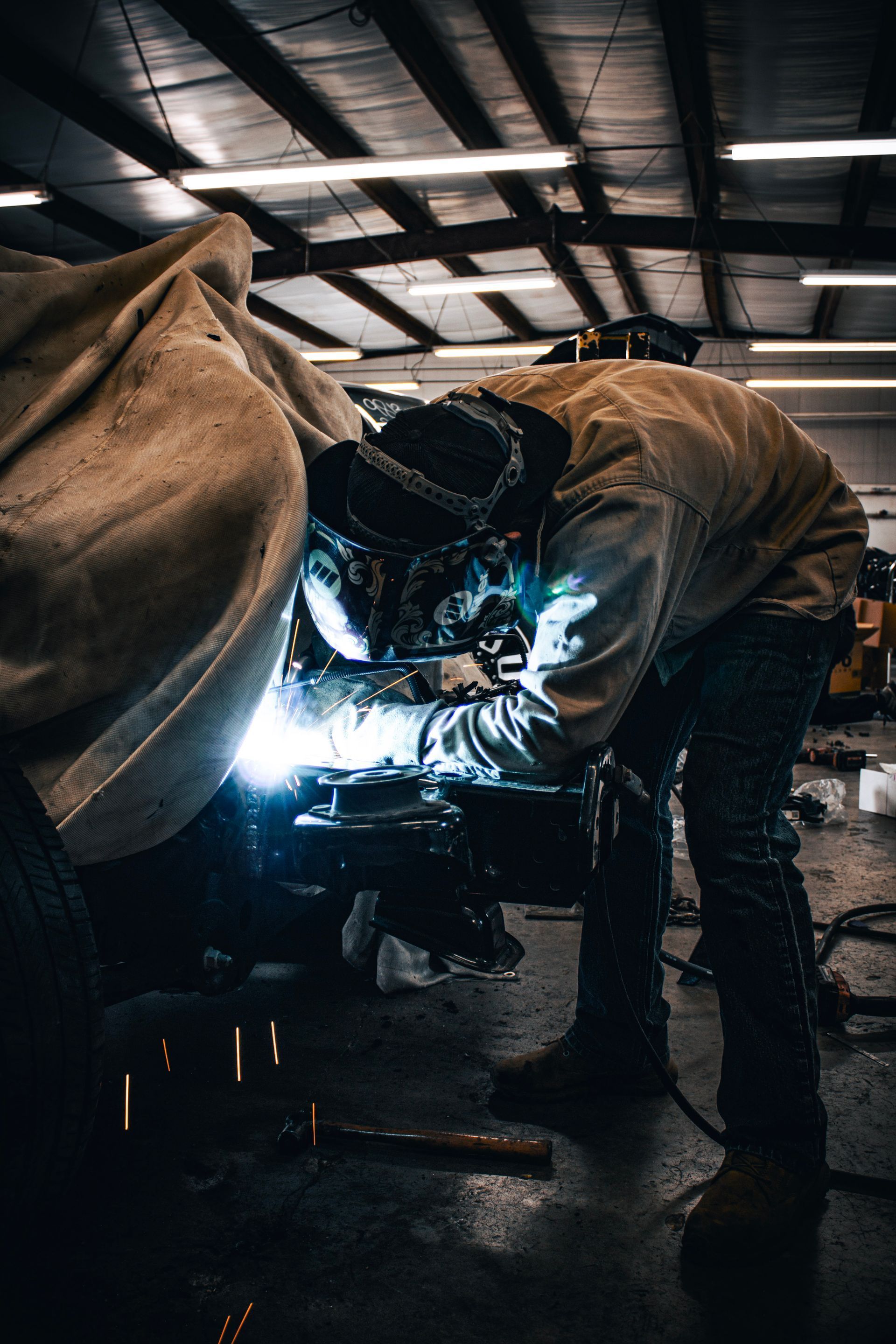 Welder working on metal in a shop, sparks flying. They are wearing protective gear, leaning over the work.