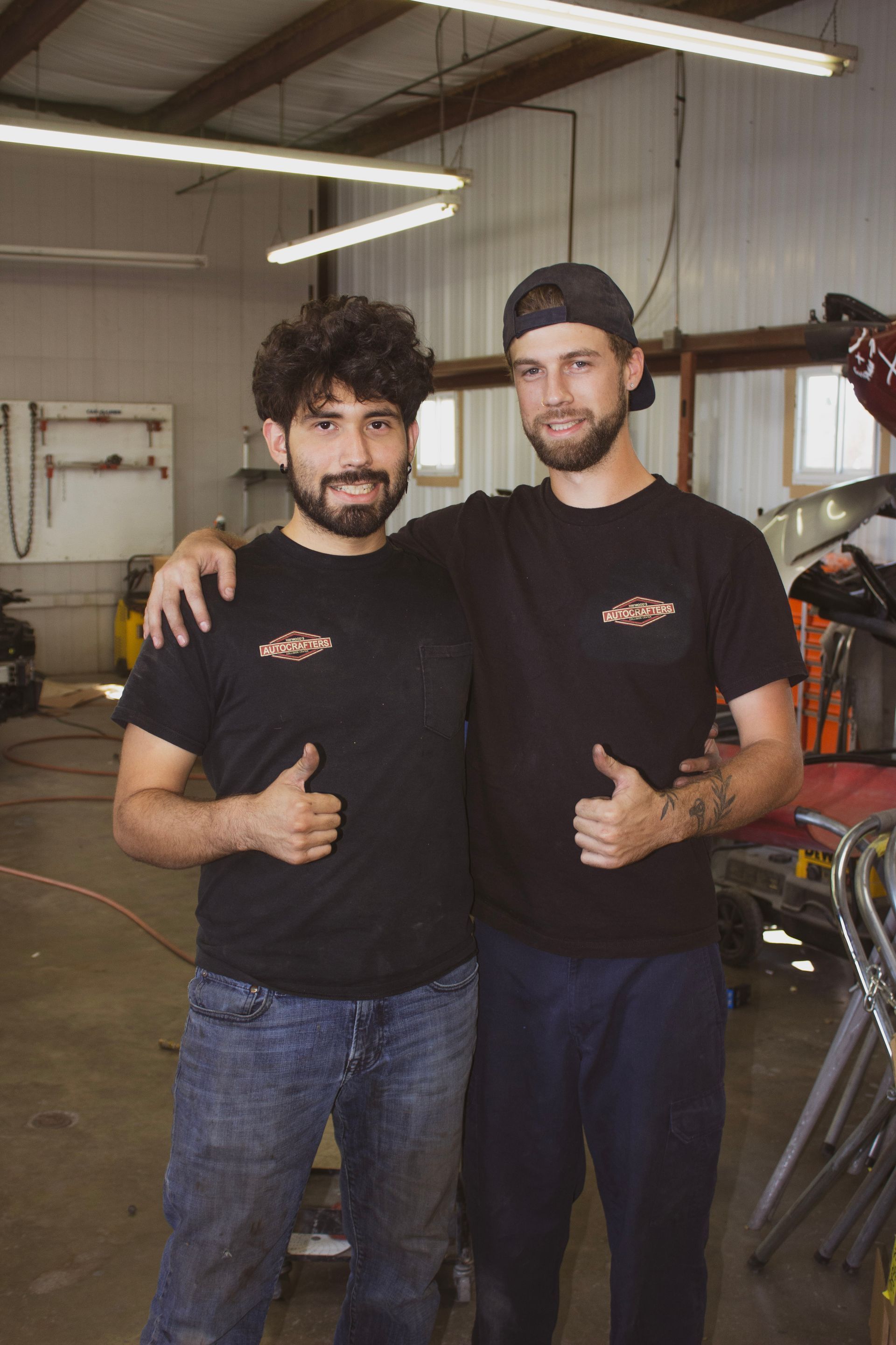 Two men in a garage with thumbs up, wearing black shirts.