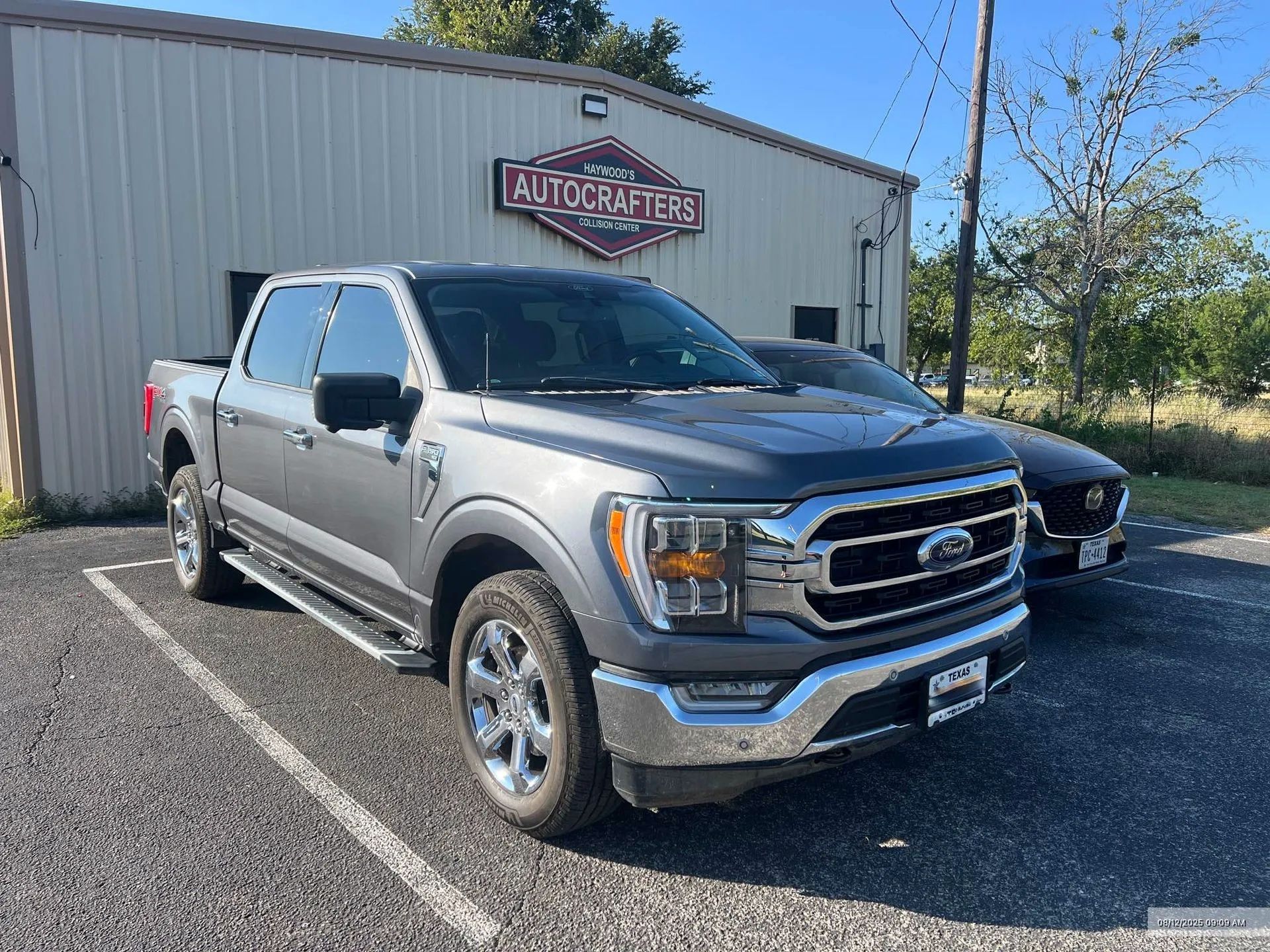 Gray Ford F-150 pickup truck parked in front of a building with a sign. A black Mustang is parked nearby.