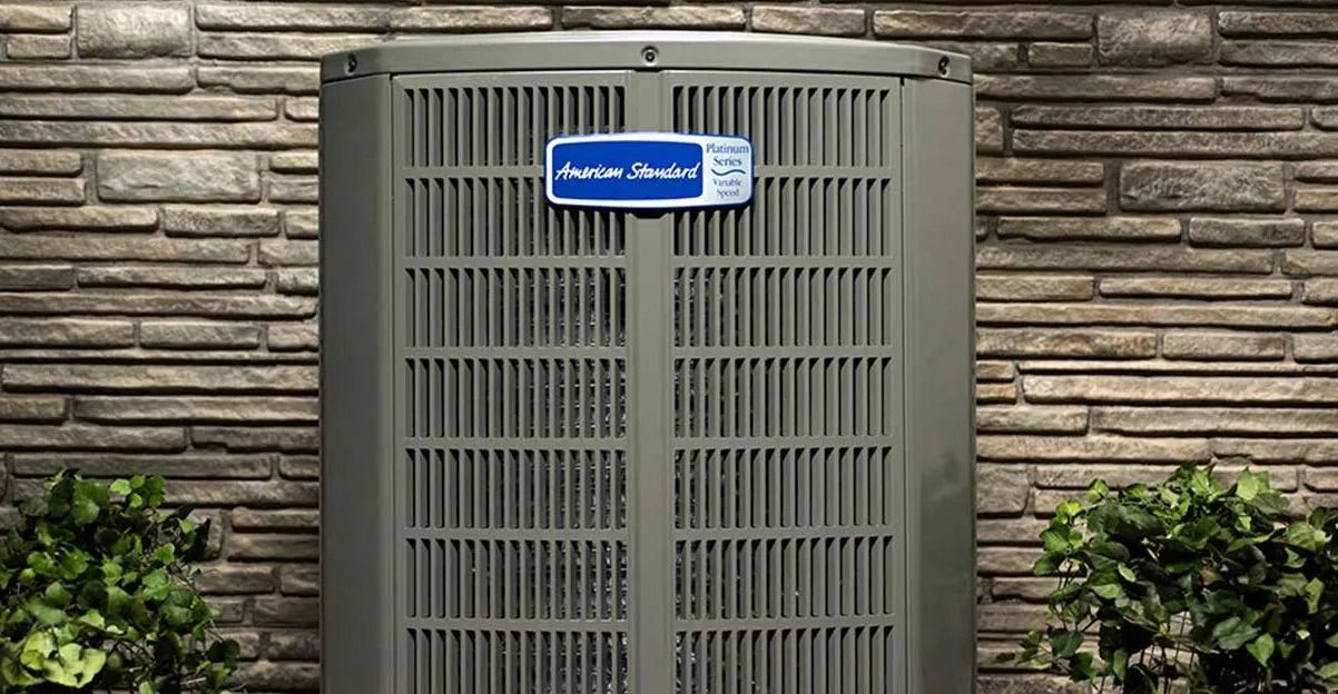 American Standard air conditioner unit against a stone wall, outdoors.