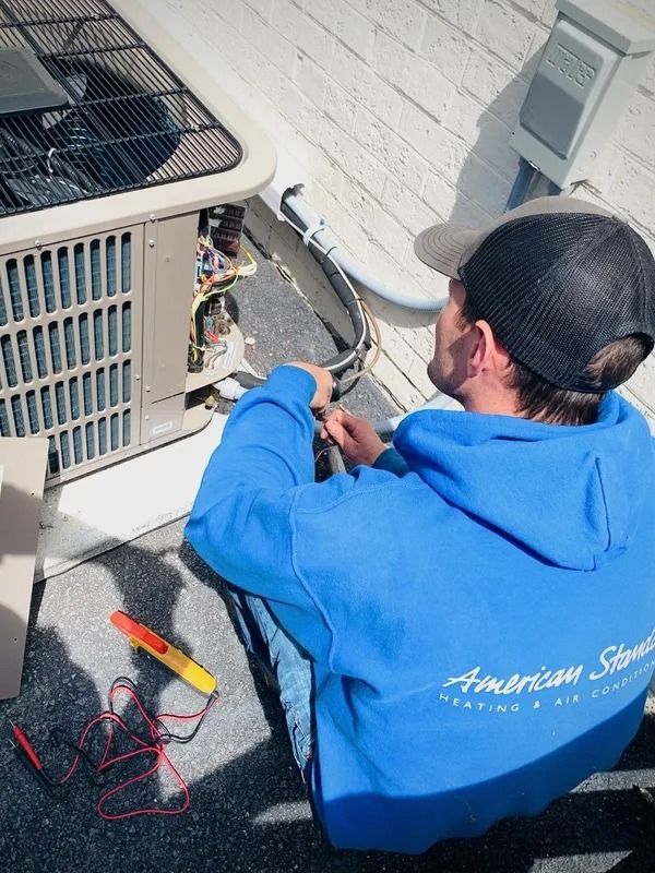 HVAC technician in blue hoodie repairs an air conditioner unit. Black cap, tools and wires are visible. Outdoors.
