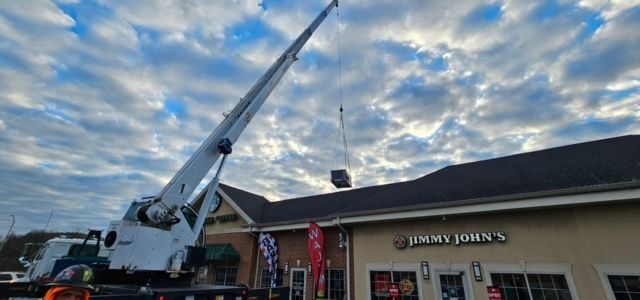 Crane lifting a dark-colored object toward the roof of a Jimmy John's restaurant under a cloudy sky.